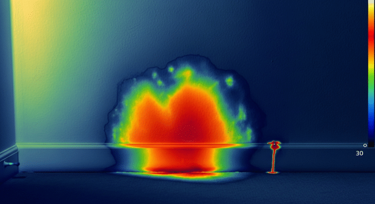Wärmebild, das eine Wasserschadenstelle an einer Innenwand in Bodennähe zeigt, mit leuchtendem Rot und Gelb, die auf hohe Temperatur oder Feuchtigkeit hinweisen, und kühleren Blau- und Grüntönen an den Rändern.
