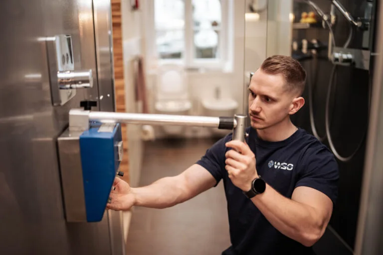 Ein Mann in einem marineblauen Hemd benutzt einen Metallhebel an einem blauen Wandgerät und führt eine Wasserschadenortung in einem modernen Badezimmer mit einer Toilette und einer Dusche im Hintergrund durch.