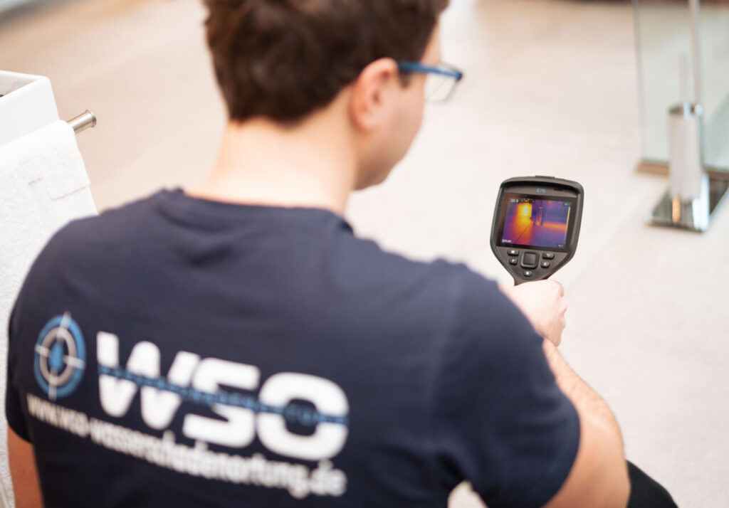 Eine Person, die eine Brille und ein marineblaues Hemd mit der Aufschrift WSO auf dem Rücken trägt, verwendet eine tragbare Wärmebildkamera zur Wasserschadenortung in Innenräumen und betrachtet einen Bildschirm mit einer Wärmekarte.