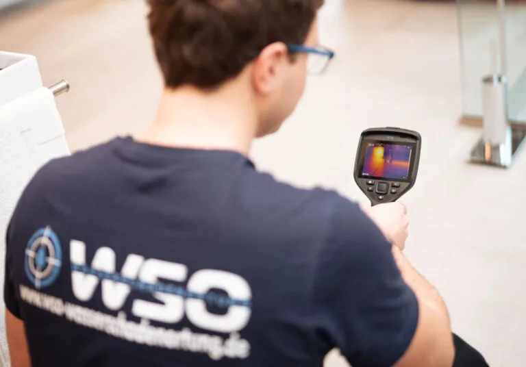 Eine Person, die eine Brille und ein marineblaues Hemd mit der Aufschrift WSO auf dem Rücken trägt, verwendet eine tragbare Wärmebildkamera zur Wasserschadenortung in Innenräumen und betrachtet einen Bildschirm mit einer Wärmekarte.