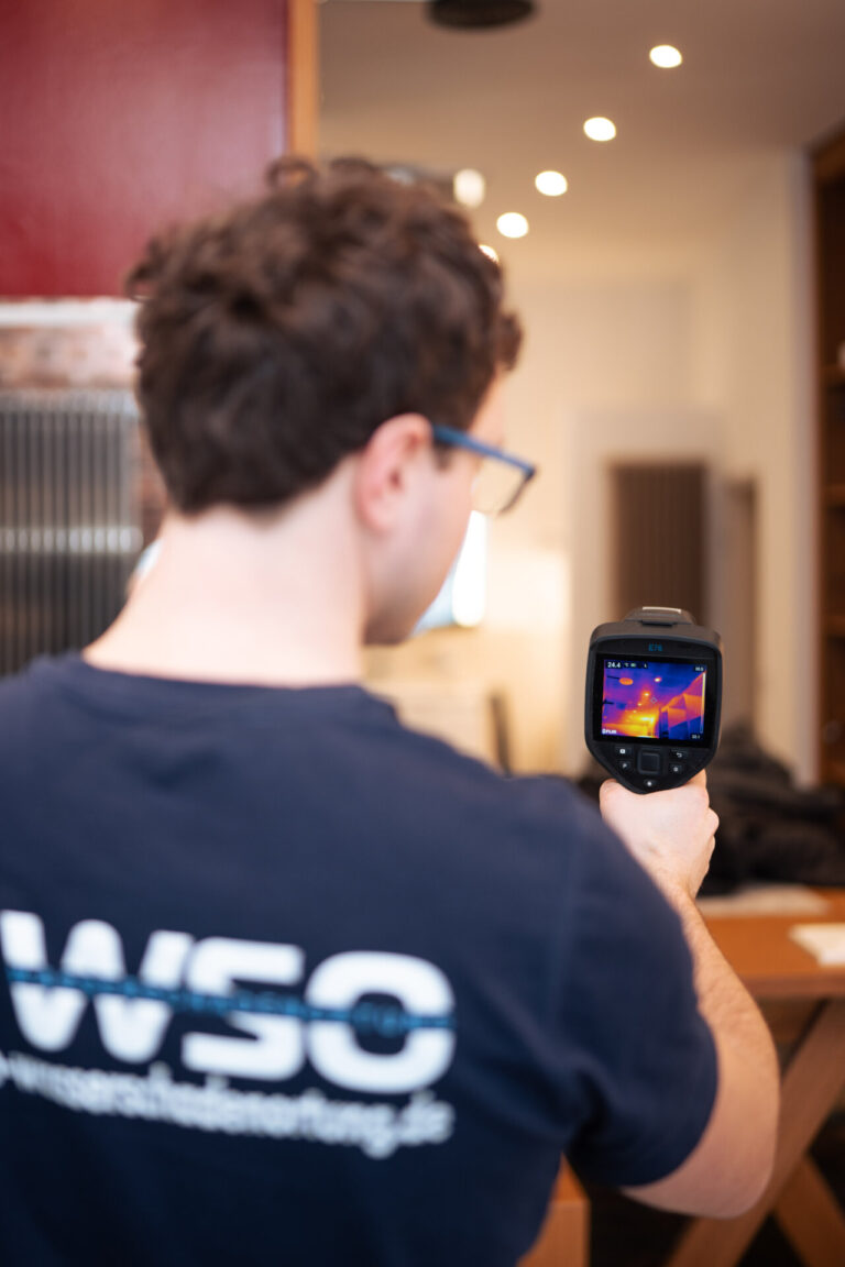 Eine Person mit kurzen braunen Haaren benutzt eine tragbare Wärmebildkamera für die Wasserschadenortung, um einen Raum auf Feuchtigkeit zu untersuchen. Auf dem Kameradisplay wird eine Wärmekarte angezeigt. Die Person trägt ein marineblaues Hemd mit weißer Schrift auf dem Rücken.