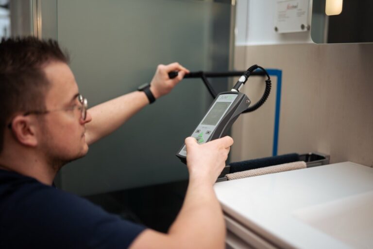 Eine Brillenträgerin benutzt ein elektronisches Handmessgerät mit einer Sonde, um die Wasserschadenortung neben einem Waschbecken und einem Spiegel durchzuführen.