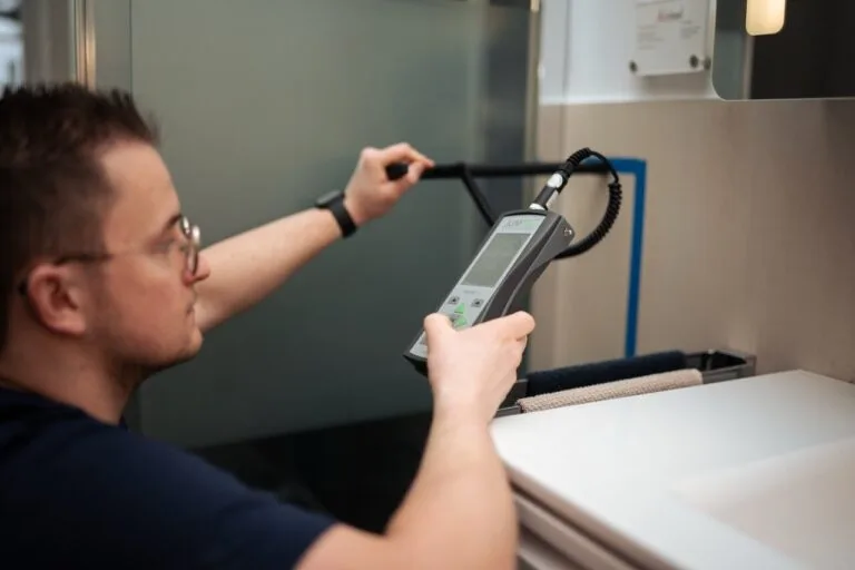 Eine Brillenträgerin benutzt ein elektronisches Handmessgerät mit einer Sonde, um die Wasserschadenortung neben einem Waschbecken und einem Spiegel durchzuführen.