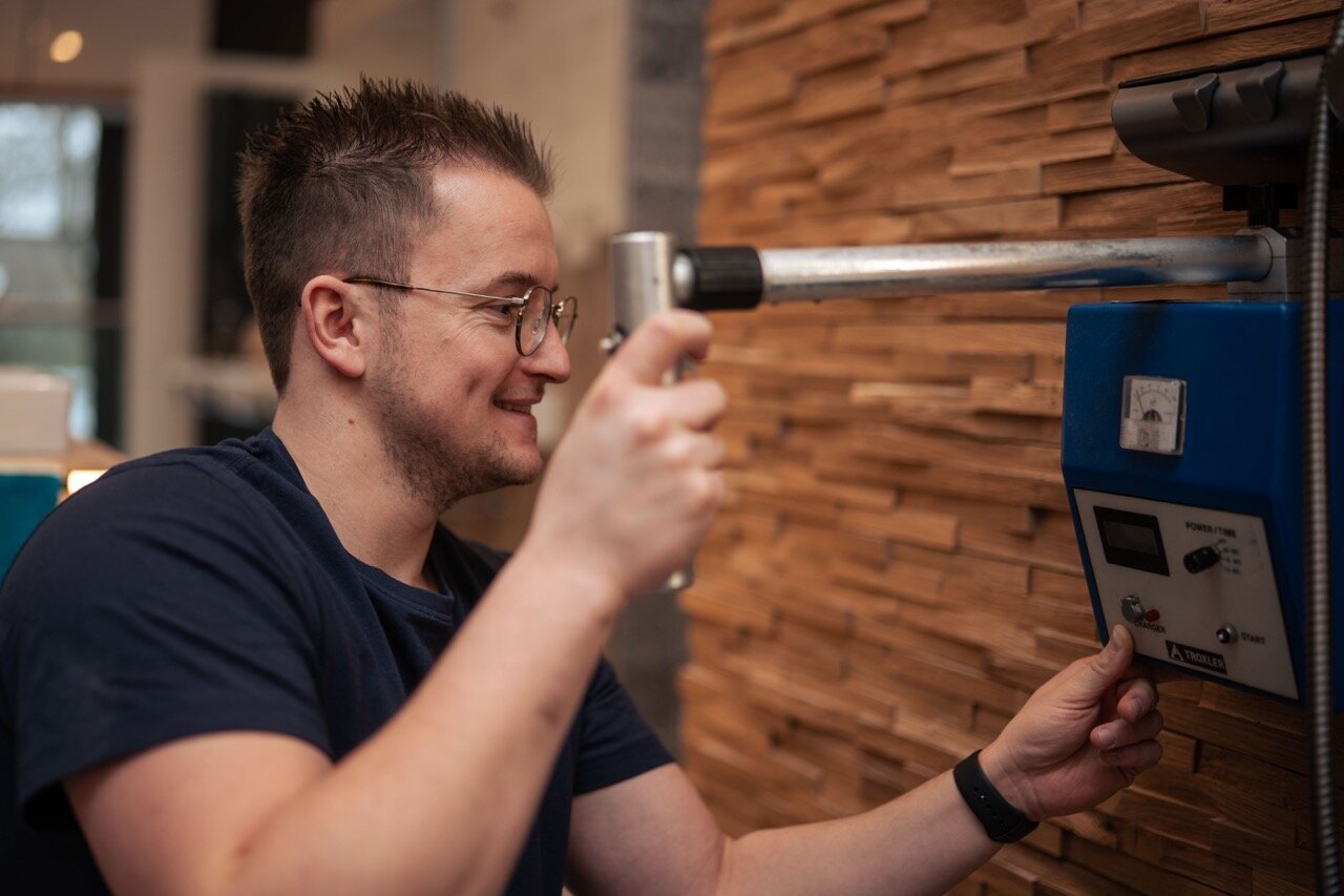 Ein Mann mit Brille lächelt, während er in einem modernen Raum mit Holzwänden eine blau-silberne Wasserschadenortungsmaschine bedient und deren Regler einstellt.