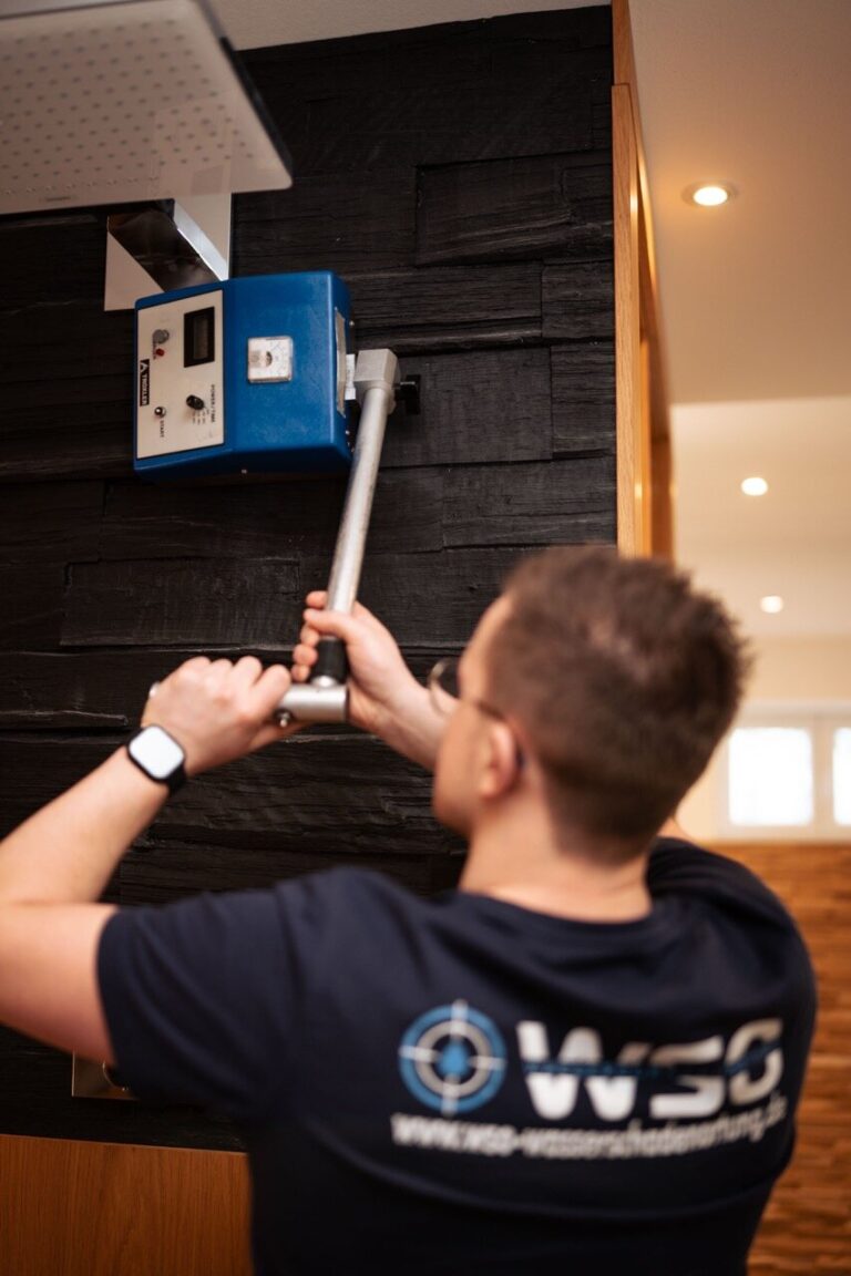Eine Person, die eine Brille und ein T-Shirt mit W3C-Logo trägt, benutzt ein Metallwerkzeug zur Wasserschadenortung, während sie ein blaues Gerät, das an einer schwarzen Wand in einem Innenraum angebracht ist, einstellt oder repariert.
