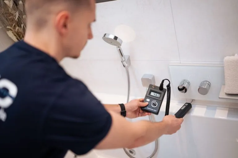 Eine Person in einem marineblauen Hemd benutzt ein digitales Gerät zur Wasserschadenortung neben dem Wasserhahn einer weißen Badewanne in einem modernen Badezimmer. Im Hintergrund sind ein Duschkopf und ein gefaltetes Handtuch zu sehen.