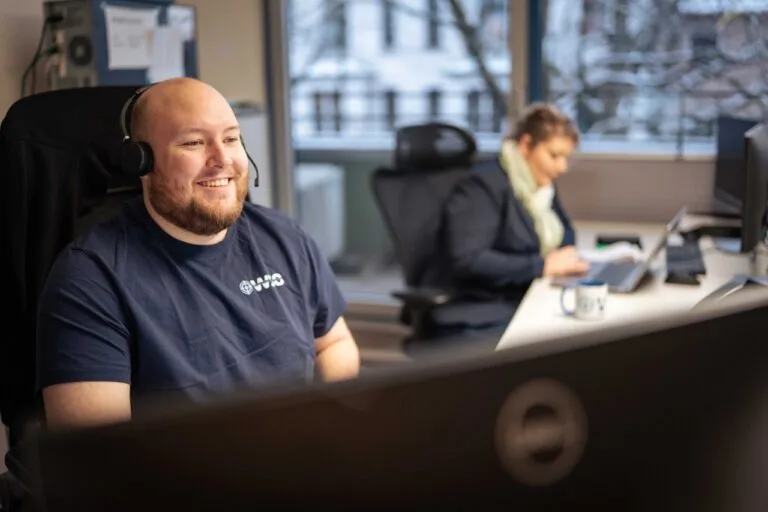 Ein l&auml;chelnder Mann, der ein Headset tr&auml;gt, sitzt an einem Schreibtisch mit einem Computermonitor im Vordergrund, w&auml;hrend eine Frau in der N&auml;he eines Fensters arbeitet, vor dem eine verschneite Landschaft liegt - ideale Teamarbeit, um Aufgaben wie die Wasserschadenortung effizient zu erledigen.