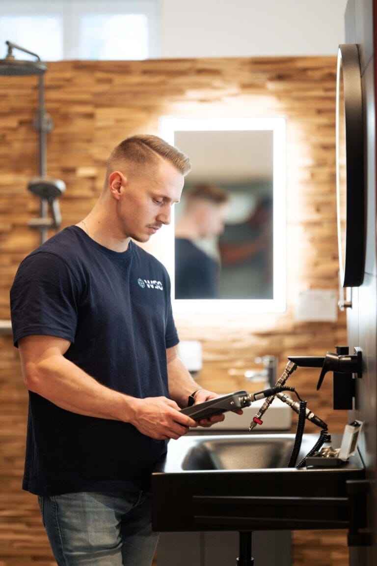 Ein Mann mit kurzen blonden Haaren steht an einem modernen Waschbecken im Badezimmer, trägt ein marineblaues T-Shirt und blickt nach unten, während er ein kleines schwarzes Wasserschadenortungsgerät hält. Im Hintergrund sind ein großer Spiegel und eine hölzerne Wanddekoration zu sehen.