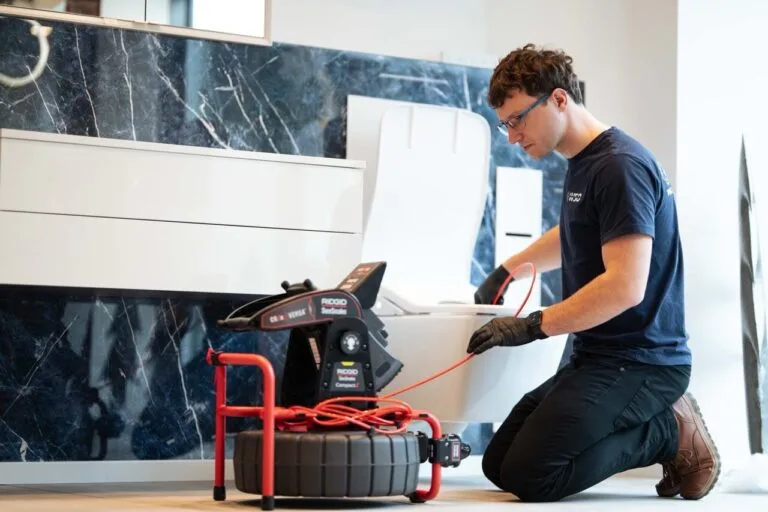Eine Person kniet mit Handschuhen auf dem Boden eines Badezimmers und bedient eine Kanalisationsinspektionskamera mit einem roten Kabel für die Wasserschadenortung, das sie zur Inspektion oder Wartung in eine Toilette führt.