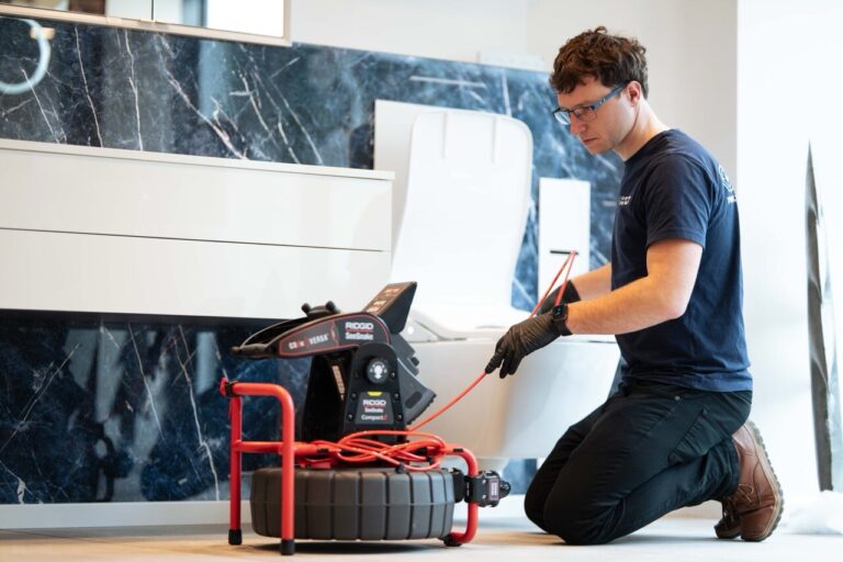 Eine Person mit Brille und Handschuhen kniet auf dem Boden und benutzt ein RIDGID-Inspektionskamerasystem in der Nähe eines Waschbeckens und einer Toilette, um möglicherweise Sanitäranlagen oder Rohre auf Wasserschadenortung zu überprüfen.