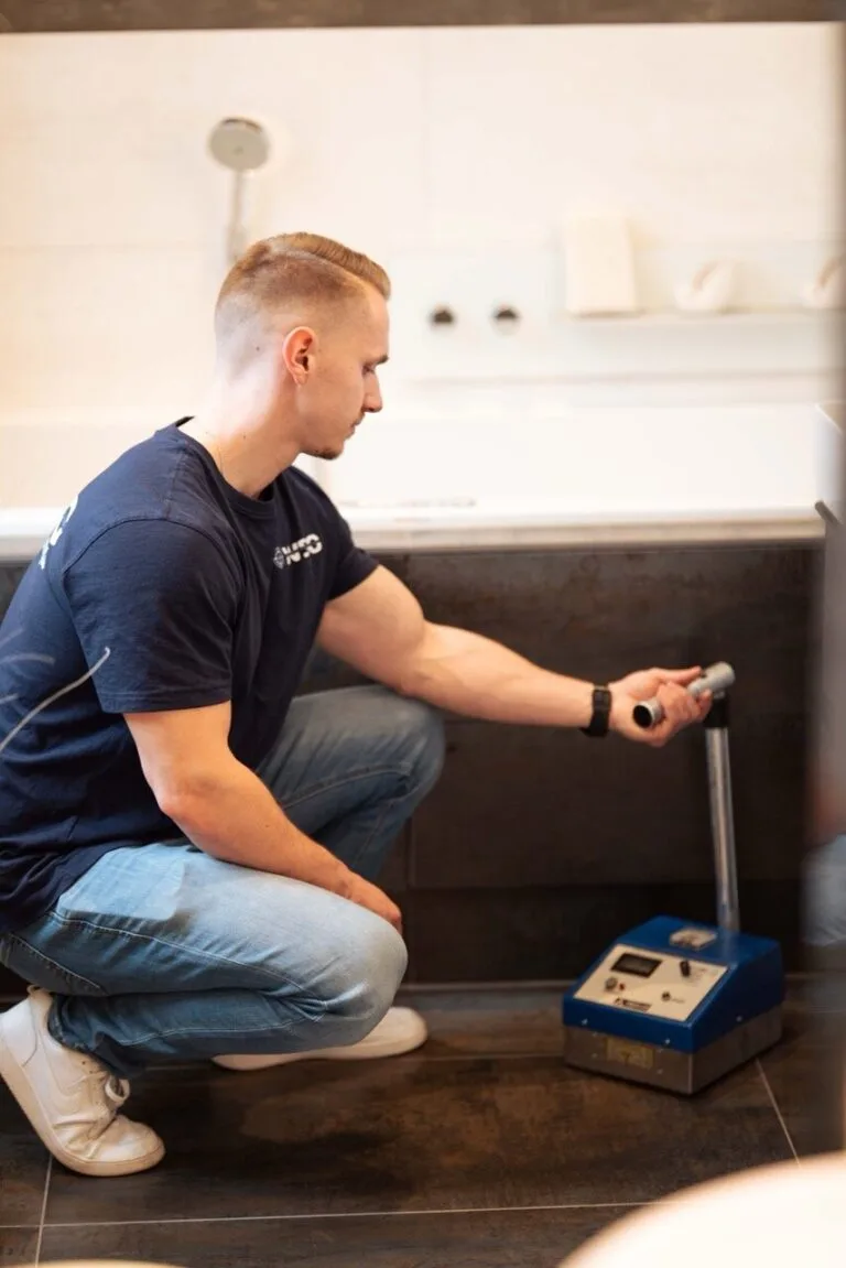 Ein Mann, der ein marineblaues Hemd und Jeans trägt, kniet neben einer Badewanne und benutzt eine Metallsonde zur Wasserschadenortung, die mit einem blauen Testgerät verbunden ist, das auf dem Boden steht.