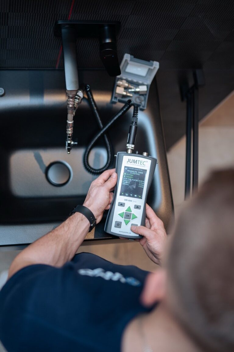Eine Person misst mit einem JUMTEC-Handgerät mit Display und Tasten in der Nähe eines schwarzen Waschbeckens und Wasserhahns - möglicherweise für die Wasserqualität, die Inspektion von Sanitäranlagen oder die Wasserschadenortung.