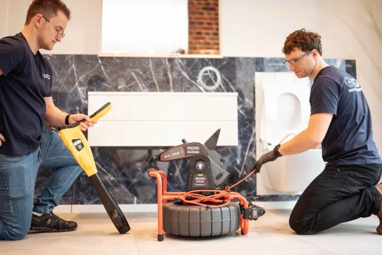 Zwei Männer in marineblauen Hemden bedienen in einem modernen Badezimmer eine Rohrinspektionskamera zur Wasserschadenortung; einer hält einen Monitor, während der andere ein orangefarbenes Kabel in das Gerät einführt. Im Hintergrund sind eine Toilette und ein Waschbecken zu sehen.