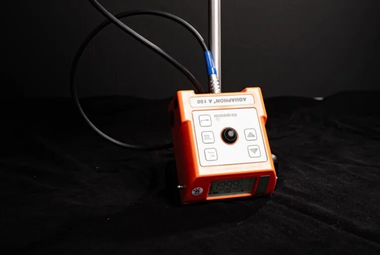 Ein orangefarbenes Aquatron A-150-Gerät mit angeschlossenen Kabeln, das für die Wasserschadenortung verwendet wird, zeigt Zahlen auf einem digitalen Bildschirm an, während es auf einer schwarzen Fläche vor einem dunklen Hintergrund steht.