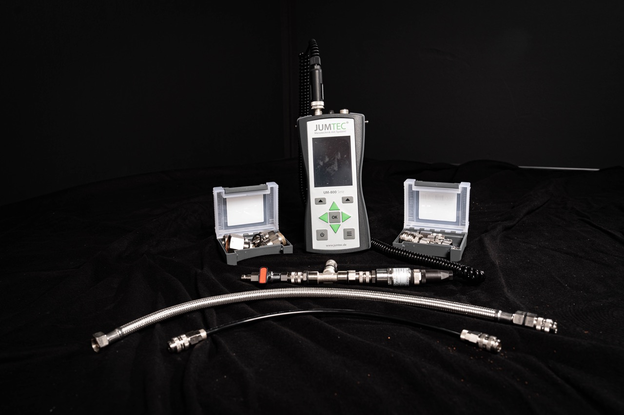 Ein JUMTEC-Handgerät für die Wasserschadenortung mit Bildschirm und Tasten ist umgeben von zwei offenen Gehäusen mit kleinen Metallteilen und zwei Metallschläuchen, die auf einem schwarzen Stoffhintergrund dargestellt sind.