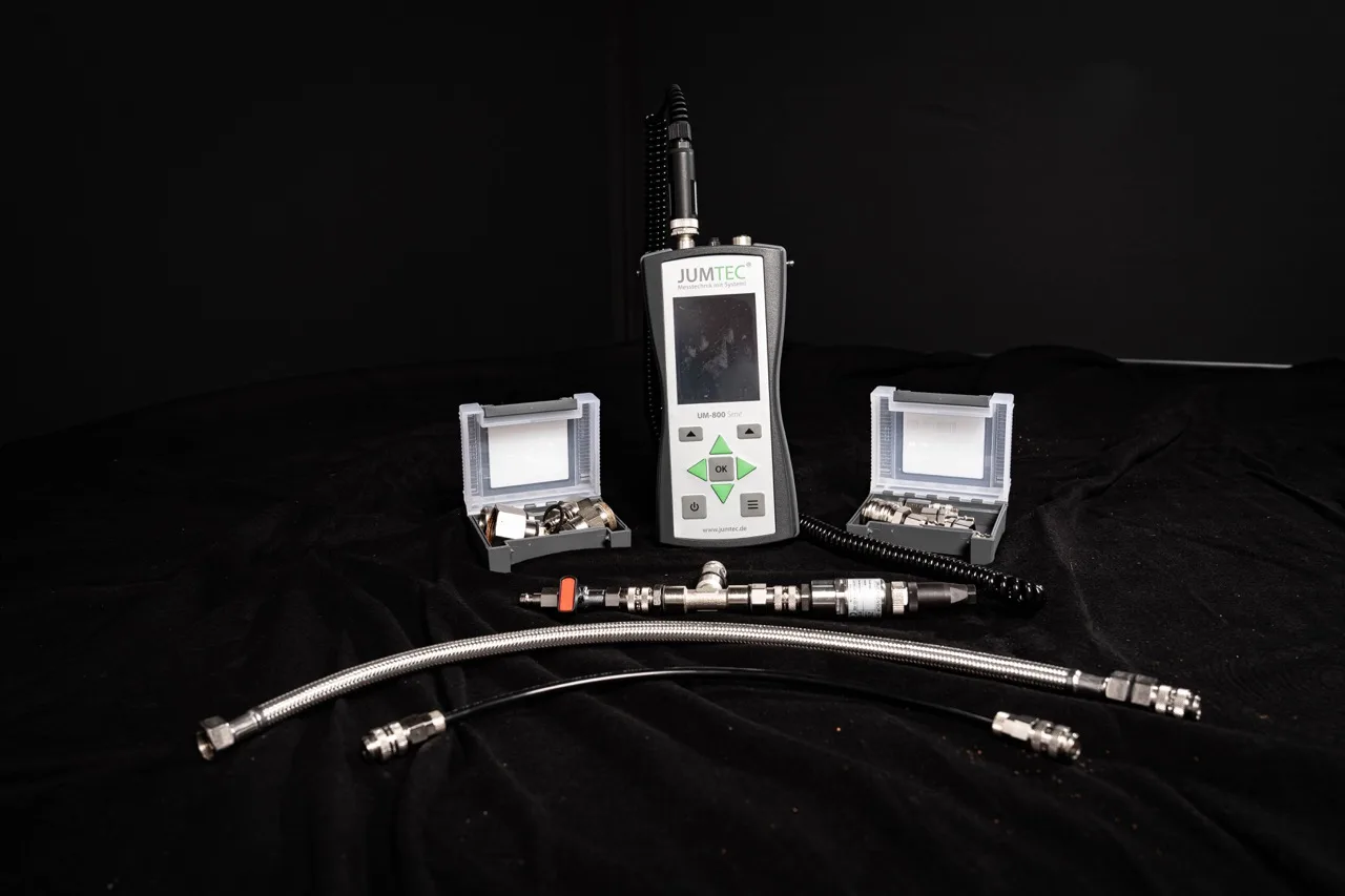 Ein JUMTEC-Handgerät für die Wasserschadenortung mit Bildschirm und Tasten ist umgeben von zwei offenen Gehäusen mit kleinen Metallteilen und zwei Metallschläuchen, die auf einem schwarzen Stoffhintergrund dargestellt sind.