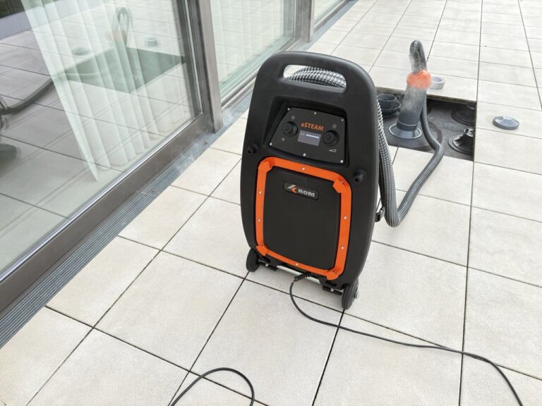 Eine schwarz-orangefarbene Dampfreinigungsmaschine mit Rädern steht auf einem gefliesten Boden neben einem Glasfenster, bereit für die Wasserschadenortung. Schläuche und Kabel sind befestigt, und im Hintergrund ist ein weiteres Reinigungsgerät zu sehen.