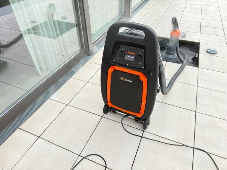 Eine schwarz-orangefarbene Dampfreinigungsmaschine mit Rädern steht auf einem gefliesten Boden neben einem Glasfenster, bereit für die Wasserschadenortung. Schläuche und Kabel sind befestigt, und im Hintergrund ist ein weiteres Reinigungsgerät zu sehen.