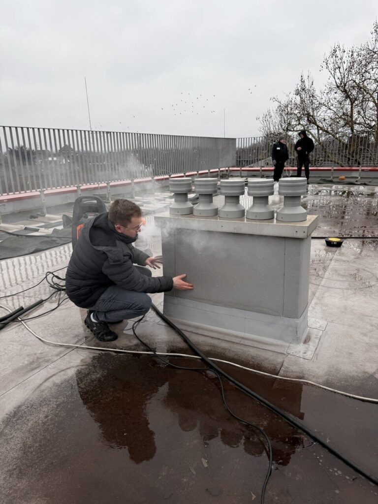 Eine Person in einer Winterjacke kniet auf einem Dach und untersucht und wärmt ihre Hände in der Nähe eines dampfenden Lüftungsschachts, möglicherweise bei der Wasserschadenortung. Zwei Personen stehen im Hintergrund an einem Geländer unter einem bedeckten Himmel.