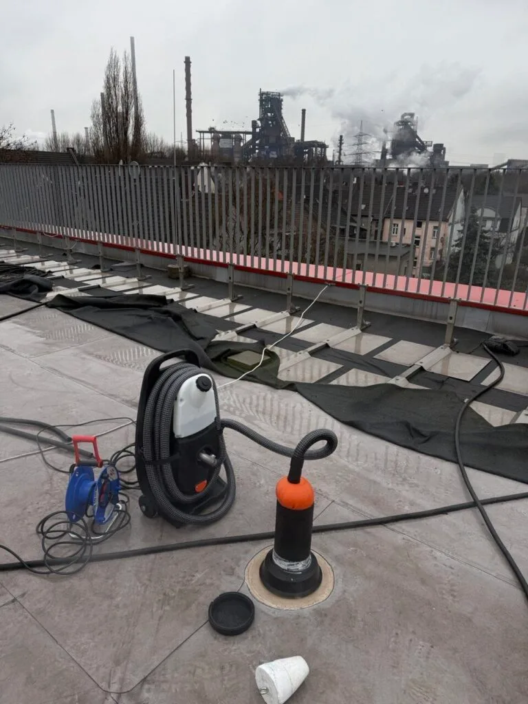 Ein Nass-/Trockensauger, eine Verlängerungskabeltrommel und eine orangefarbene Tauchpumpe für die Wasserschadenortung stehen auf einem Flachdach mit freiliegendem Abdichtungsmaterial. Im Hintergrund raucht an einem bewölkten Tag ein Stahlwerk.