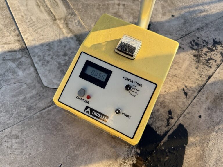 Nahaufnahme eines gelben Trokler-Geräts, das für die Wasserschadenortung auf einer Betonfläche verwendet wird und auf seinem digitalen Bildschirm 00:55 anzeigt, mit Beschriftungen für Ladegerät, Start, Strom-/Zeiteinstellungen und einem Metallteil obenauf.