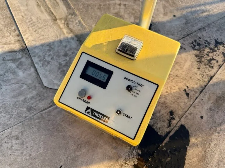 Nahaufnahme eines gelben Trokler-Geräts, das für die Wasserschadenortung auf einer Betonfläche verwendet wird und auf seinem digitalen Bildschirm 00:55 anzeigt, mit Beschriftungen für Ladegerät, Start, Strom-/Zeiteinstellungen und einem Metallteil obenauf.