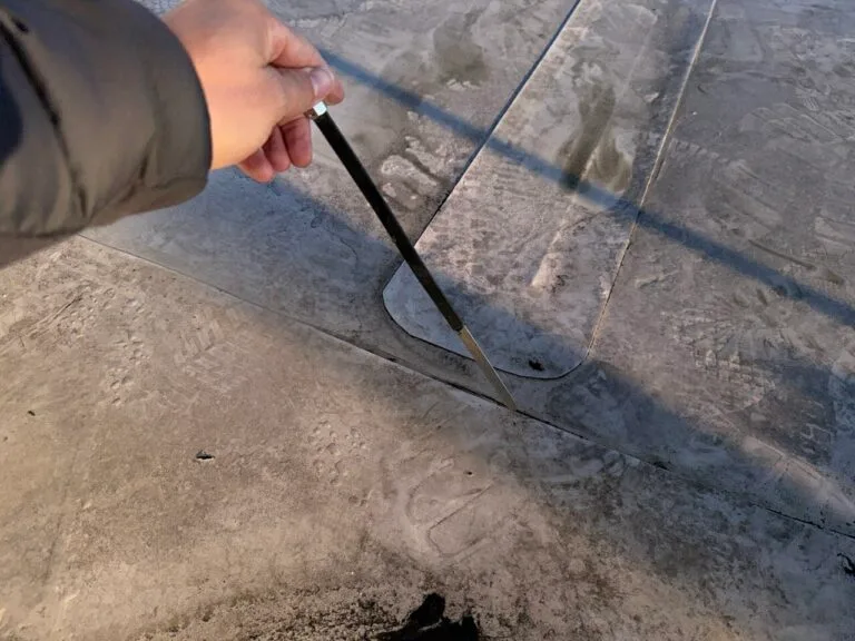 Eine Hand, die einen Metallstab hält, zeigt auf einen schmalen Spalt in einer staubigen, mit Fußspuren versehenen Betonfläche - eine typische Inspektionsszene während der Wasserschadenortung.