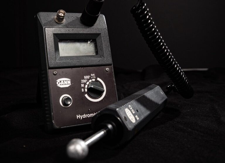 Nahaufnahme eines GANN Hydromette-Feuchtigkeitsmessgeräts, das für die Wasserschadenortung unerlässlich ist, mit Digitalanzeige, Zifferblatt und angebrachter Sonde vor einem dunklen Hintergrund.