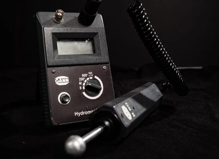 Nahaufnahme eines GANN Hydromette-Feuchtigkeitsmessgeräts, das für die Wasserschadenortung unerlässlich ist, mit Digitalanzeige, Zifferblatt und angebrachter Sonde vor einem dunklen Hintergrund.