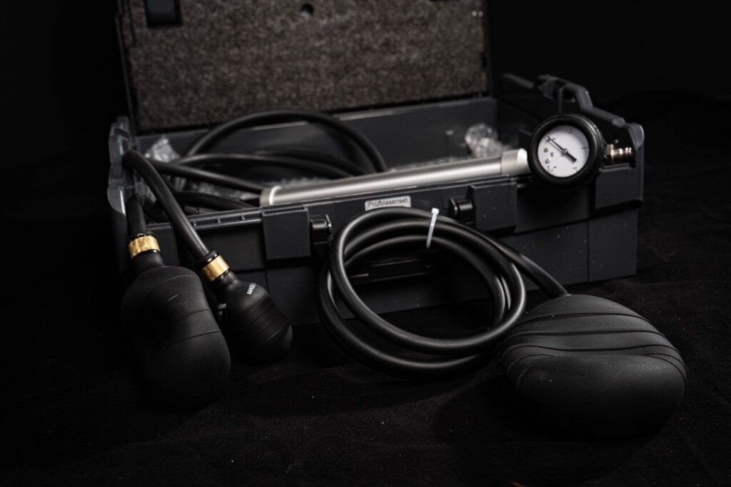 Ein schwarzer Kunststoffkoffer, der ein Manometer, zwei schwarze Gummilampen mit Schläuchen und zusätzliche Schläuche für eine präzise Wasserschadenortung enthält, alles vor einem dunklen Hintergrund.
