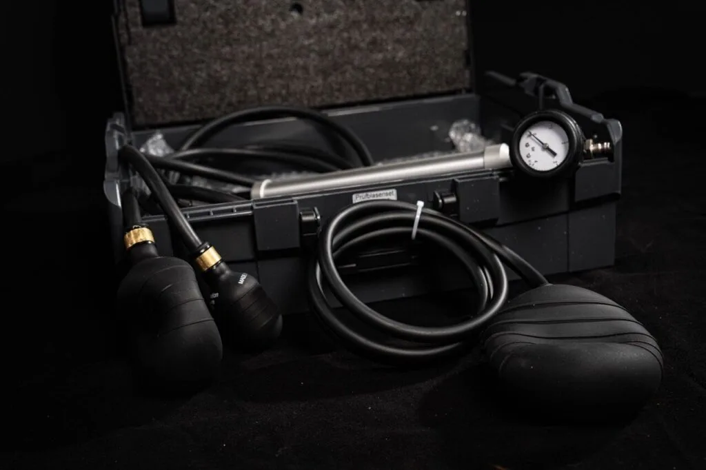 Ein schwarzer Kunststoffkoffer, der ein Manometer, zwei schwarze Gummilampen mit Schläuchen und zusätzliche Schläuche für eine präzise Wasserschadenortung enthält, alles vor einem dunklen Hintergrund.
