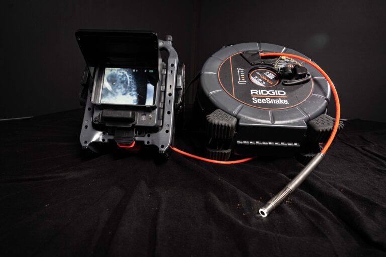Ein RIDGID SeeSnake Kamerasystem, ideal für die Wasserschadenortung, mit einem Bildschirm, der das Rohrinnere zeigt, ist an einem langen flexiblen Kabel befestigt und sitzt auf einer schwarzen Fläche.