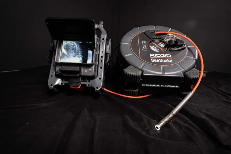 Ein RIDGID SeeSnake Kamerasystem, ideal für die Wasserschadenortung, mit einem Bildschirm, der das Rohrinnere zeigt, ist an einem langen flexiblen Kabel befestigt und sitzt auf einer schwarzen Fläche.