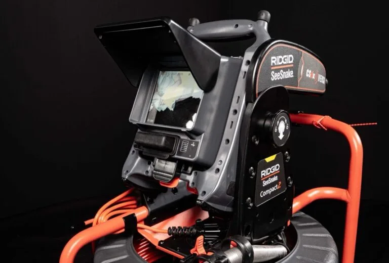 Eine Nahaufnahme eines RIDGID SeeSnake Compact2 Inspektionskamerasystems, ideal für die Wasserschadenortung, mit einem Monitor, der ein Bild anzeigt, aufgerollten orangen Kabeln und einem dunklen Hintergrund.