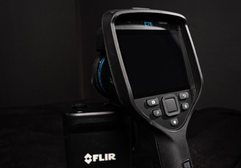 Eine Nahaufnahme einer FLIR E76-Wärmebildkamera mit schwarzem Gehäuse, das angewinkelt ist, um den Bildschirm und die Bedientasten vor einem dunklen Hintergrund zu zeigen - ideal für präzise Wasserschadenortungsanwendungen.