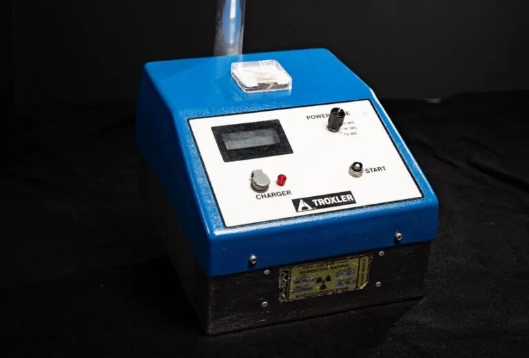 Ein blau-schwarzes elektronisches Gerät mit der Aufschrift TROXLER, das für die Wasserschadenortung verwendet wird, verfügt über eine digitale Anzeige, einen Netz- und einen Startschalter, eine Ladeanzeige und einen Griff an der Oberseite, der sich vor einem dunklen Hintergrund befindet.