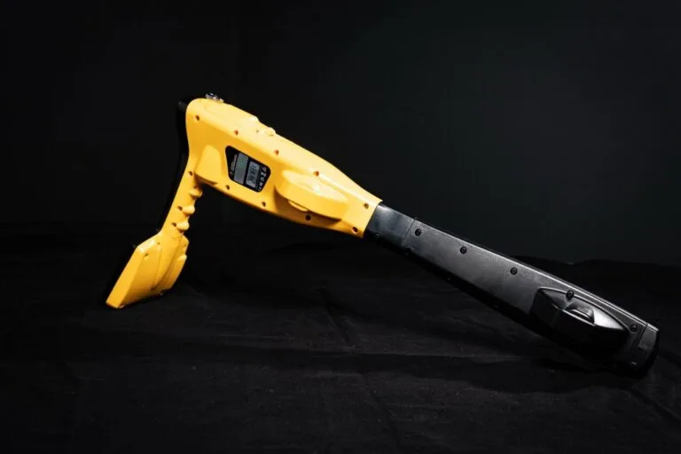 Ein gelb-schwarzer Handheld-Metalldetektor für die Wasserschadenortung steht auf einer schwarzen Fläche mit dunklem Hintergrund und zeigt seinen Bildschirm und seinen ergonomischen Griff.