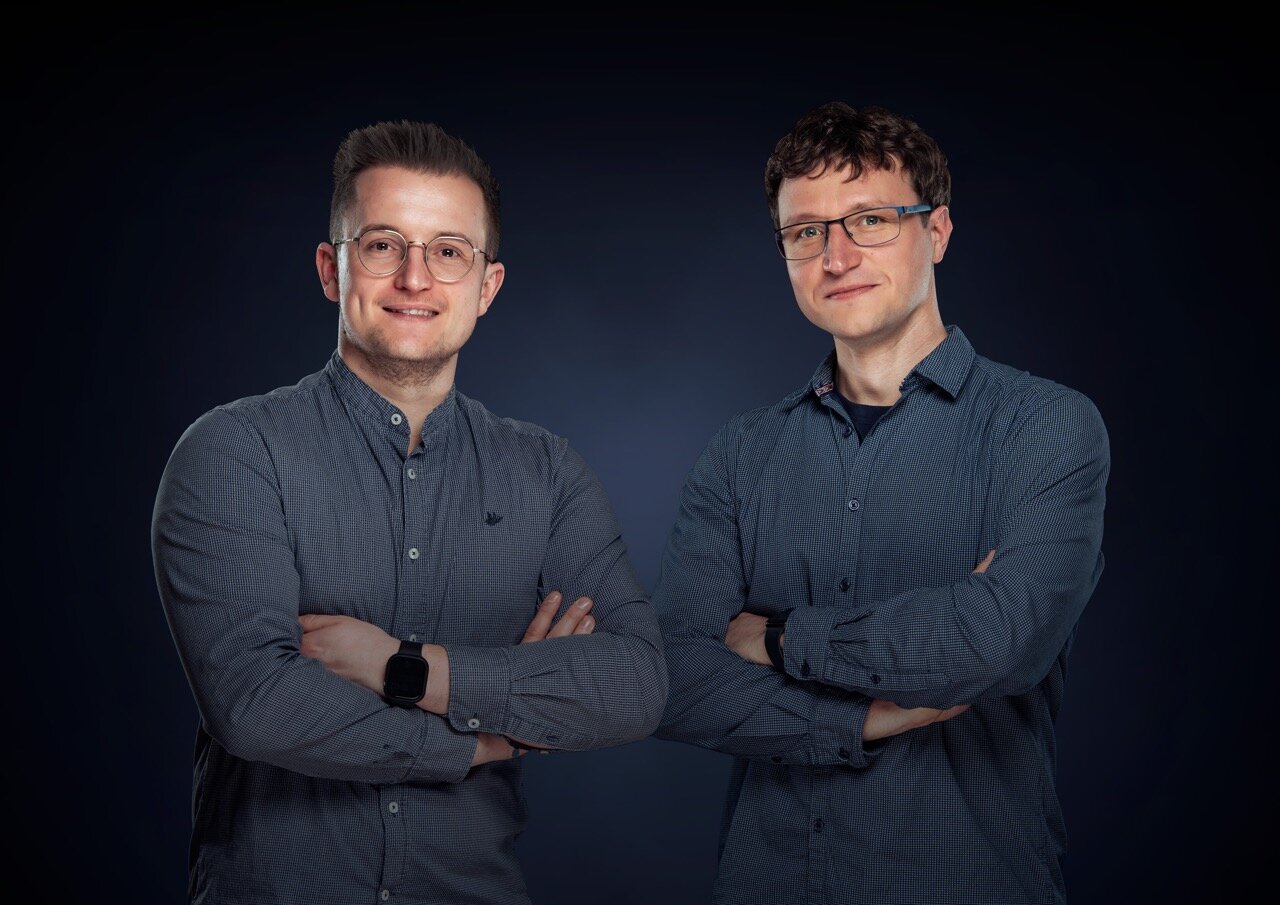 Zwei Männer mit Brillen und blauen Button-up-Hemden stehen mit verschränkten Armen nebeneinander und lächeln vor einem dunkelblauen Hintergrund in die Kamera, bereit, bei der Wasserschadenortung zu helfen.