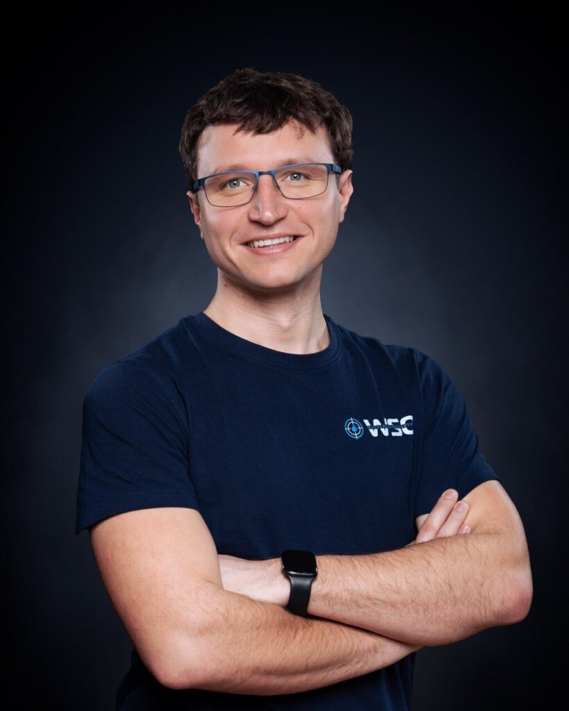 Ein lächelnder Mann mit kurzen braunen Haaren und Brille steht mit verschränkten Armen, trägt ein marineblaues T-Shirt mit einem Wasserschadenortung-Logo und eine Smartwatch vor einem dunklen, schlichten Hintergrund.