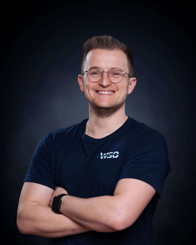 Eine lächelnde Person mit kurzen Haaren und Brille steht mit verschränkten Armen in einem marineblauen "WSO"-T-Shirt - perfekt für einen Wasserschadenortung-Profi - vor einem dunklen Hintergrund mit Farbverlauf.