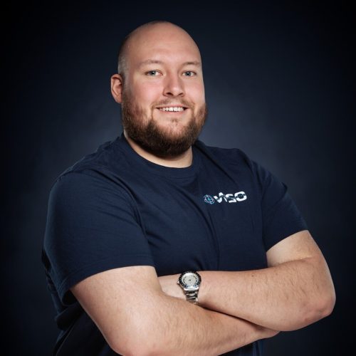 Ein glatzköpfiger Mann mit Bart, der ein marineblaues Wasserschadenortung-T-Shirt mit einem Logo und einer Armbanduhr trägt, steht lächelnd mit verschränkten Armen vor einem dunklen Hintergrund.