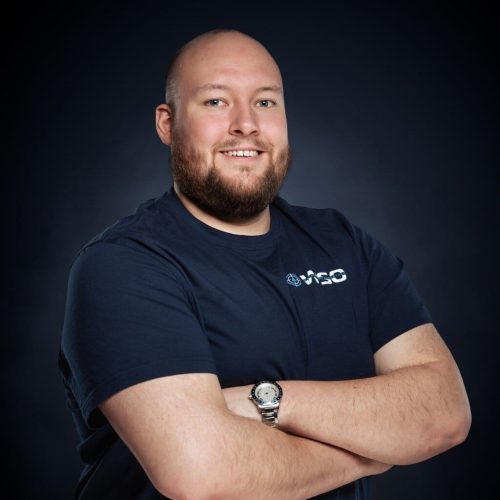 Ein glatzköpfiger Mann mit Bart, der ein marineblaues Wasserschadenortung-T-Shirt mit einem Logo und einer Armbanduhr trägt, steht lächelnd mit verschränkten Armen vor einem dunklen Hintergrund.