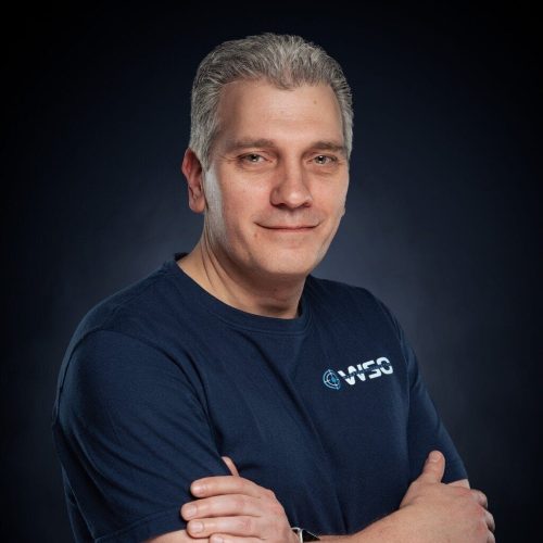 Ein Mann mittleren Alters mit kurzen grauen Haaren lächelt leicht und hat die Arme verschränkt. Er trägt ein marineblaues T-Shirt mit einem kleinen Wasserschadenortung-Logo und den Buchstaben WSO auf der Brust vor einem dunklen Hintergrund.