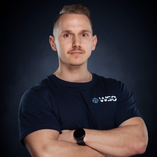 Ein Mann mit kurzen hellbraunen Haaren und einem Schnurrbart steht selbstbewusst mit verschränkten Armen, trägt ein dunkelblaues WSO Wasserschadenortung T-Shirt und eine schwarze Smartwatch vor einem dunklen Hintergrund mit Farbverlauf.