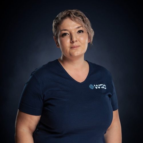 Eine Person mit kurzen, gewellten hellbraunen Haaren, die ein marineblaues Hemd mit weißem NEO-Logo trägt, steht vor einem Hintergrund mit dunklem Farbverlauf und schaut ruhig in die Kamera - bereit, bei der fachkundigen Wasserschadenortung zu helfen.